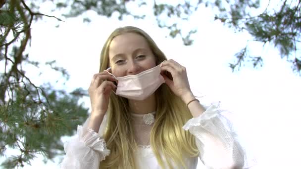 Ξανθιά νεαρή ενήλικη κοπέλα που φοράει λευκό φόρεμα χαμογελώντας και φορώντας τη μάσκα προσώπου της — Αρχείο Βίντεο