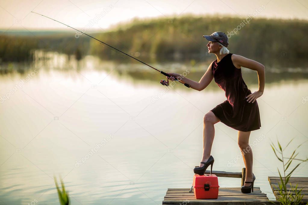 Pretty woman in a burgundy dress and black shoes and a baseball cap catches fish on a wooden pier in the spring in good weather