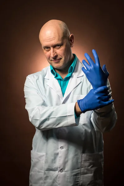 Médico experimentado de 50 años usando un abrigo blanco, usando guantes de goma azul y sonriendo Imágenes de stock libres de derechos