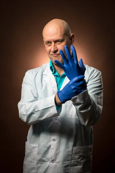 Médico experimentado de 50 años usando un abrigo blanco, usando guantes de goma azul y sonriendo Imagen de archivo