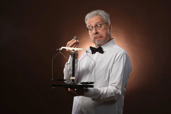 Anciano hombre de pelo gris de 50 años, en camisa blanca, gafas y pajarita que pesa algo en escamas con pesas Fotos de stock