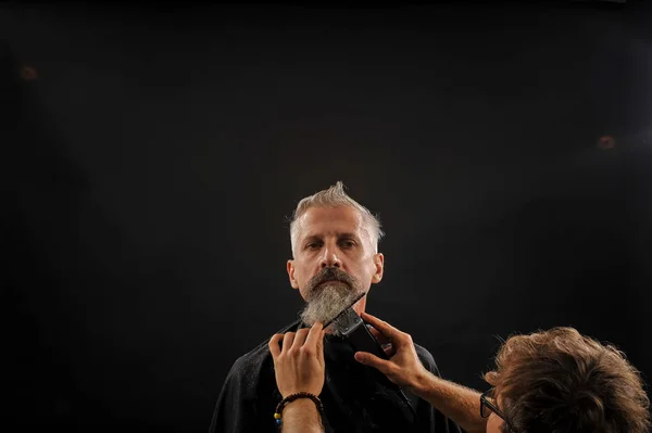 Barbero corta una barba a un cliente a un anciano canoso Imagen de archivo