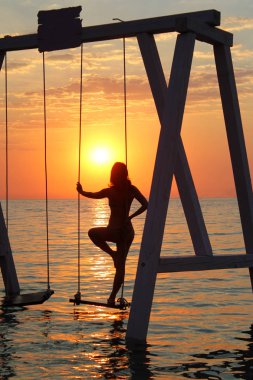 genç bir kadın denizde bir salıncak üzerinde duruyor ve şafak karşılar.