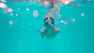 Havuzun içinden 5 yaşındaki bir çocuk yüzme gözlükleriyle dalıyor. Güneşli, mavi bulutlu bir günde yaz mevsimindeyiz. Mutlu çocuk havuzda eğleniyor..