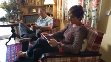 İzleme görüntüsü yaklaşıyor. Rahat yaşlı çift rahat bir oturma odasında mobil cihazlarına bakıyor. Büyükanne ve büyükbaba yaşam tarzı, gündüz.