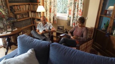 Üst düzey çiftler, büyük ebeveynler, eski koltuklarda oturup cep telefonu ve tablete bakıyorlar. Kırsal ve iyi dekore edilmiş bir evin oturma odasında. İzleme görüntüsü.