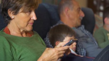 Kanepede oturan beyaz orta yaşlı büyükanne ailenin geri kalanı televizyon izlerken cep telefonuna bakıyor. Kapatın. Gündüz.