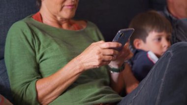 Kanepede oturan Kafkasyalı son sınıf öğrencisi ailenin geri kalanı televizyon izlerken cep telefonuna bakıyor. Kapatın. Gündüz.