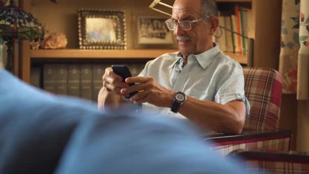 70代の白人男性で携帯電話を使って椅子に座っていました 彼は眼鏡をかけ居心地の良いダイニングルームにいる — ストック動画