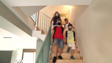 Büyük beyaz bir aileden dört çocuk, okulun ilk günü evden merdivenlerden aşağı iniyorlar. Koruyucu maskeler ve sırt çantaları takıyorlar. Yeni kurallar, COVID-19 salgını sırasındaki yeni normallik. İzleme görüntüsü.  