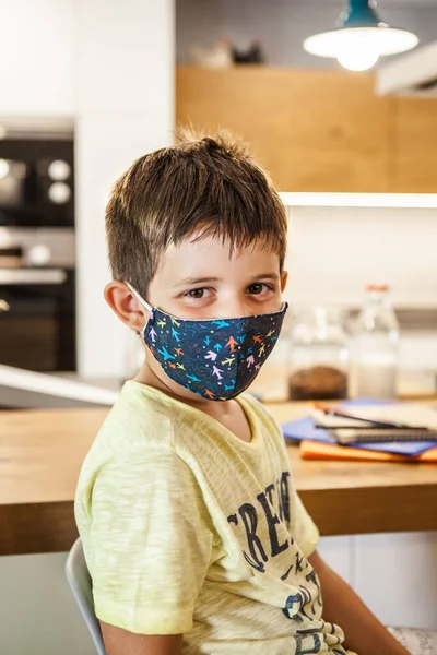 Portrait of 6-years-old caucasian boy with colored protective mask looking at camera. Preschool Kid in house kitchen. Daytime, first day of school after coronavirus pandemic. New rules Covid-19.