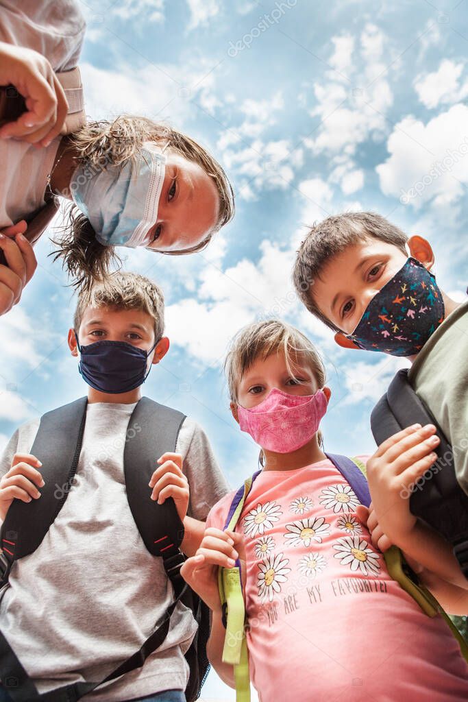 Low angle view of Children wearing protective face masks and backpacks. They are in circle looking at the camera. Cloudy blue sky behind. Concept Back to school after coronavirus.