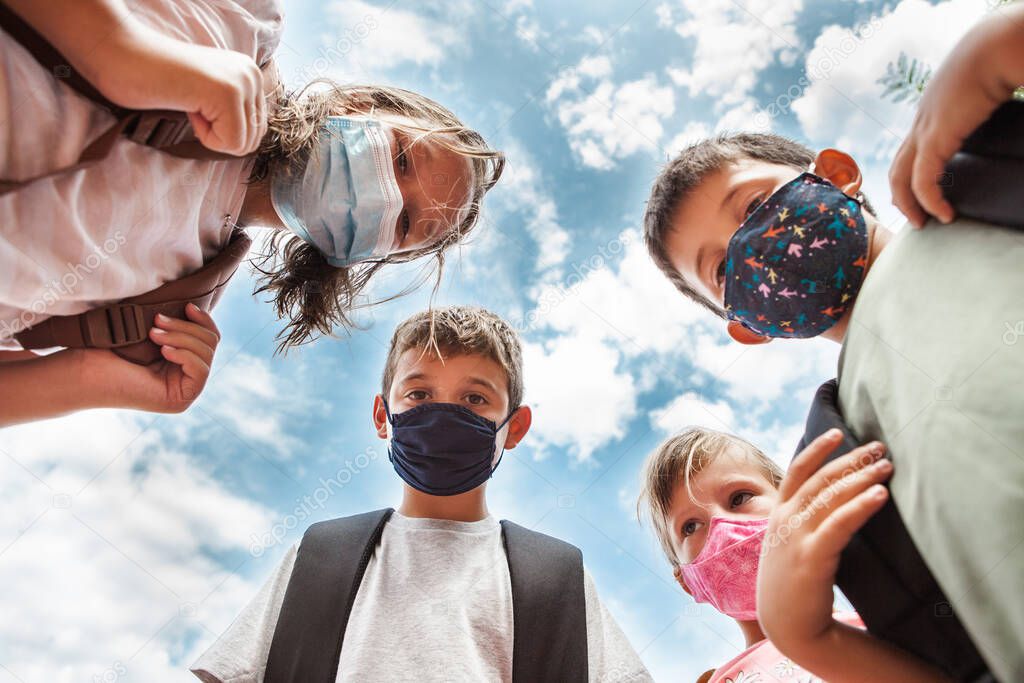 Low angle view of four children looking down at the camera, wearing colored face protective masks. They are together in circle with cloudy blue sky behind them.