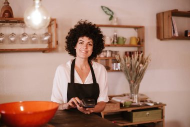 Gülümseyen kadın kafe sahibi kameraya bakıyor. Önlüklü bir kadın tezgahın arkasında duruyor ve bir kart terminalinde çalışıyor. Küçük işletme sahibi, genç girişimci kavramı.