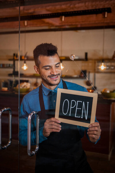 Portrait of smiling owner standing at his restaurant gate with open signboard. Young entrepreneur leaning at the cafeteria door. Chef or waiter standing in front of coffee shop.