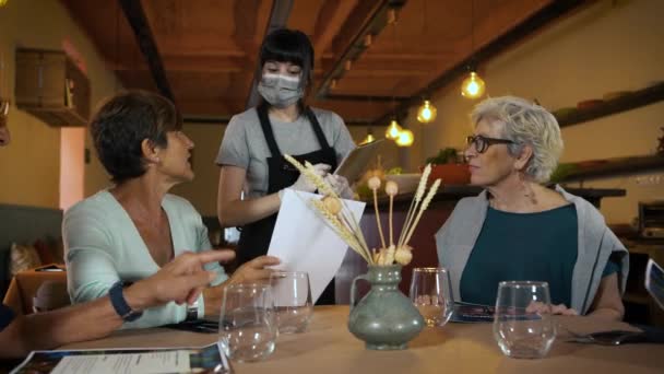 レストランで料理を注文する手にメニューを持つ2人の幸せな先輩女性 保護顔マスクと手袋を注文を取って若いウェイトレス コロナウイルス発生中の食品や飲み物 — ストック動画