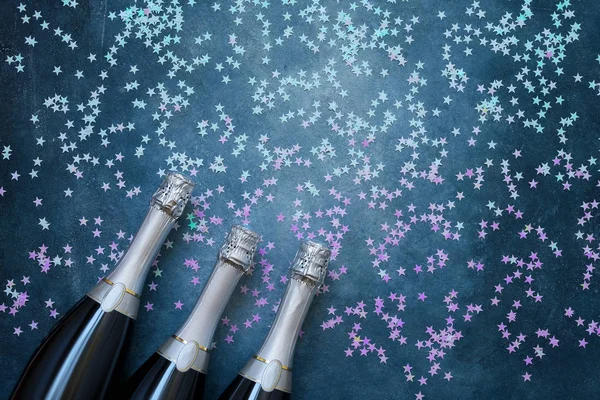 Tres botellas de champán con estrellas holográficas de confeti sobre fondo azul. Copiar espacio, vista superior — Foto de Stock