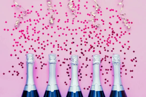 Cinco botellas de champán con estrellas de confeti y serpentinas de fiesta sobre fondo rosa. Copiar espacio, vista superior . — Foto de Stock