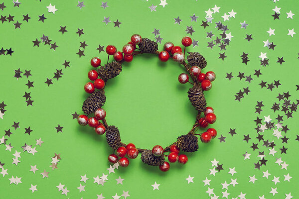 Small Christmas wreath and silver confetti stars on green background. Copy space, top view