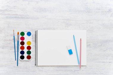 Kalem, boya ve fırçalar ile beyaz ahşap arka plan üzerinde Sanatçının boş eskiz defteri. Sanatsal maket şablonu. Üst görünüm, kopyalama alanı. Okul konseptine geri dön.