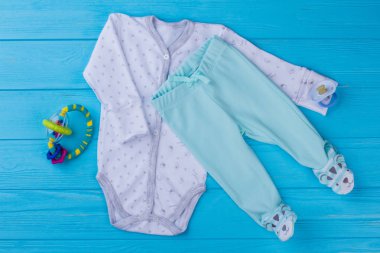 Bebek pijama ve rattle oyuncak. Beyaz toddler onesie ve komik ayaklı pantolon. Mavi ahşap arka plan.