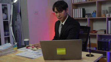 Genç bir işadamı, geceleri bir ofiste çalışırken, belgelerle ve teknolojiyle çevrili dizüstü bilgisayarına odaklanmış bir belge buluyor..