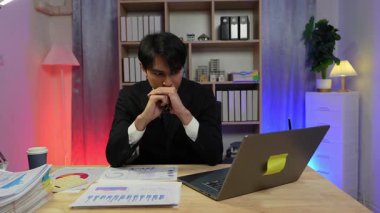 Genç bir işadamı, geceleri bir ofiste çalışırken, belgelerle ve teknolojiyle çevrili dizüstü bilgisayarına odaklanmış bir belge buluyor..