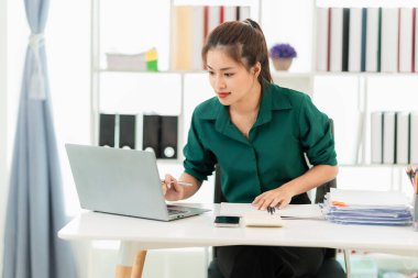 Masada doküman ve dizüstü bilgisayarla çalışan genç Asyalı kadın muhasebeci, sonuçtan tatmin olmuş, hesap makinesi kullanıyor ve vergi formlarını kontrol ediyor..