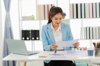Masada doküman ve dizüstü bilgisayarla çalışan genç Asyalı kadın muhasebeci, sonuçtan tatmin olmuş, hesap makinesi kullanıyor ve vergi formlarını kontrol ediyor..