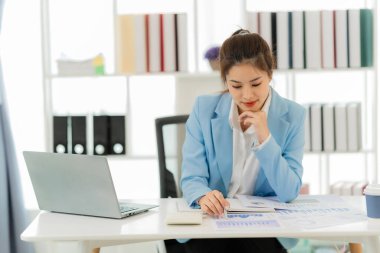 Masada doküman ve dizüstü bilgisayarla çalışan genç Asyalı kadın muhasebeci, sonuçtan tatmin olmuş, hesap makinesi kullanıyor ve vergi formlarını kontrol ediyor..