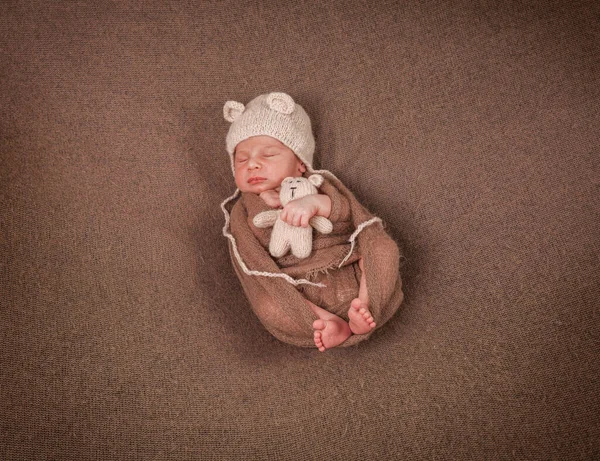 戴着一顶有耳朵的帽子 手里拿着一只棕色背景的泰迪熊的新生儿 — 图库照片