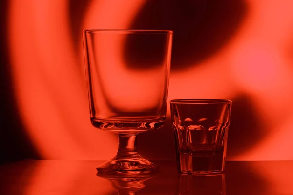Transparent large wine glass and a small stack on the background of red backlight. The concept of different possibilities