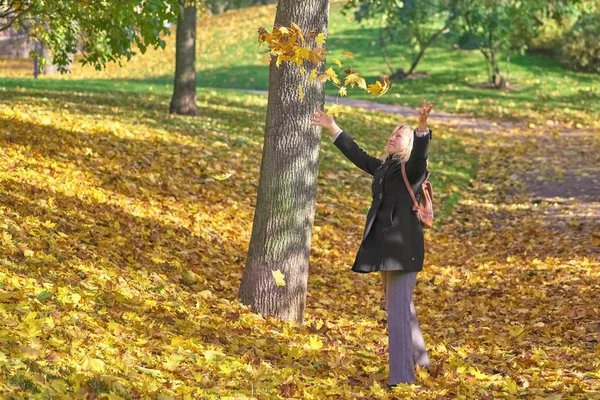 Rubia de mediana edad en un parque de otoño arroja hojas caídas a la —  Fotos de Stock