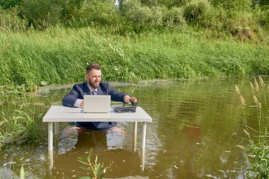 Young businessman sits waist-deep in a swamp at an office desk clipart