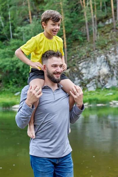 El padre lleva sobre sus hombros a su hijo a lo largo del río. Suma Imágenes De Stock Sin Royalties Gratis