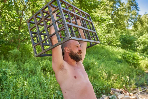 Un hombre barbudo con torsos desnudos sostiene una jaula de hierro sobre su cabeza — Foto de Stock