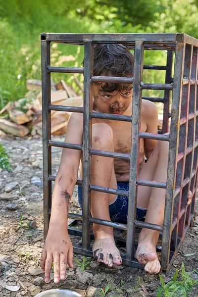 Un niño sucio y desvestido se sienta en una jaula de hierro afuera, con un gran ángulo Imagen De Stock