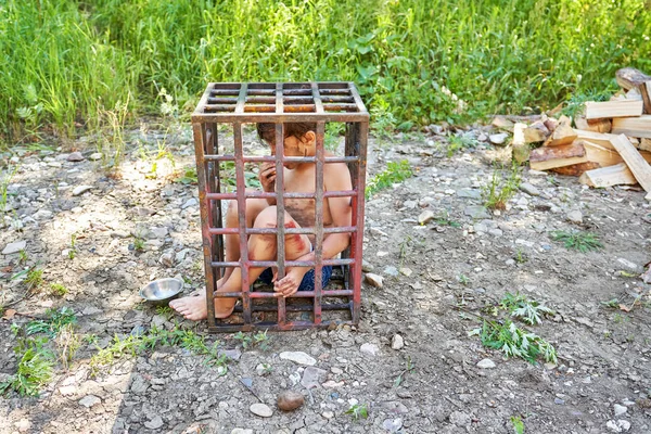 Un niño sucio se planta en una jaula de hierro al aire libre Fotos De Stock Sin Royalties Gratis