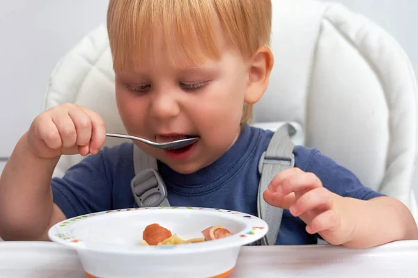 Il bimbo dai capelli rossi mangia con un cucchiaio da un piatto da bambini — Foto Stock