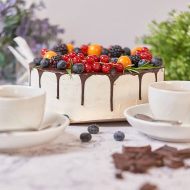 Yuvarlak beyaz pasta, koyu çikolataya bulanmış ve taze böğürtlenlerle süslenmiş. Beyaz çay fincanları ve çiçeklerle dolu bir hayat. Kare kare