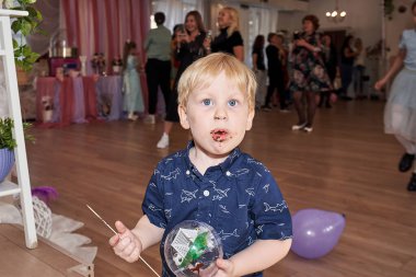Küçük bir çocuk yemek salonunun arka planında çikolatayla ağzını kirletti. Elinde oyuncak tutan bir çocuk.