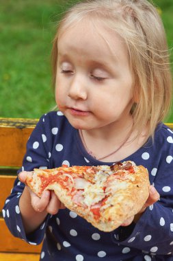 Üç yaşında, gözleri kapalı bir kız elinde bir dilim pizza tutuyor. Sokaktaki yiyecekler, dikey çerçeve, yakın çekim.