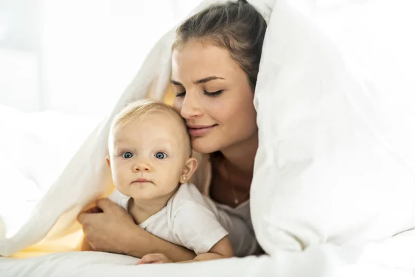 Retrato de jovem mãe com bebê menina deitada na cama coberta com um cobertor branco — Fotografia de Stock