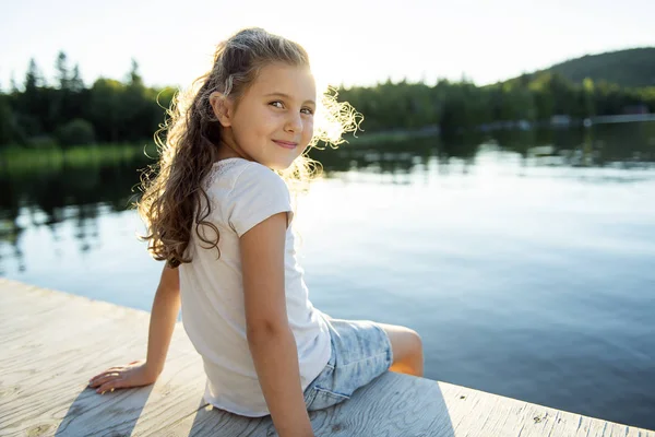 Söta barn flicka sitter på en plattform av trä vid sjön. — Stockfoto