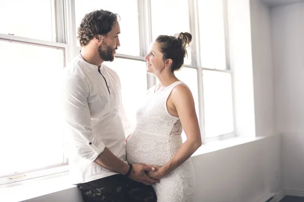 Hombre guapo y su hermosa esposa embarazada de pie cerca de la ventana en casa — Foto de Stock