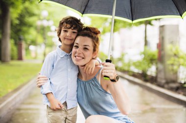 mother and child on a rainy day in a park with umbrella clipart