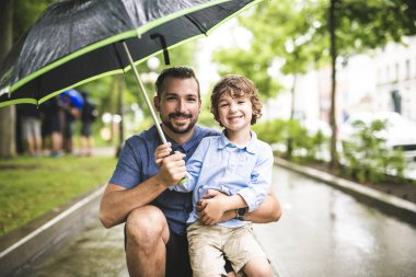 Father and child on a rainy day in a park with umbrella clipart