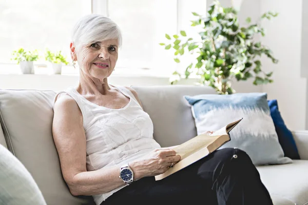 Äldre kvinna läser bok hemma vid spisen — Stockfoto