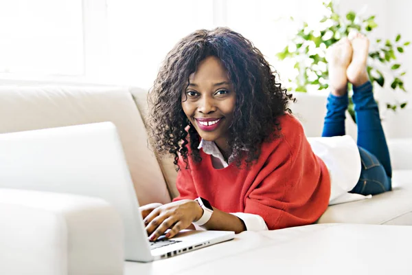 Ελκυστική μαύρη νεαρή γυναίκα χρησιμοποιώντας ένα φορητό υπολογιστή ενώ χαλαρώνετε στον καναπέ στο σπίτι — Φωτογραφία Αρχείου