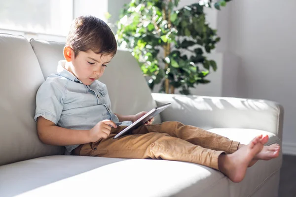 Menino com tablet digital sentado no sofá, em casa fundo interior — Fotografia de Stock